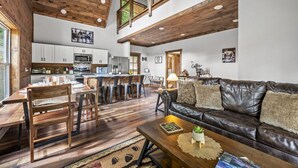 A spacious living area with a high ceiling, wooden beams, a leather sofa, a dining table with chairs, and a kitchen in the background.
