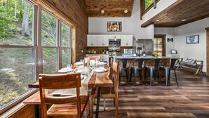 A rustic cabin interior with a dining area, kitchen, and large windows overlooking a forest.