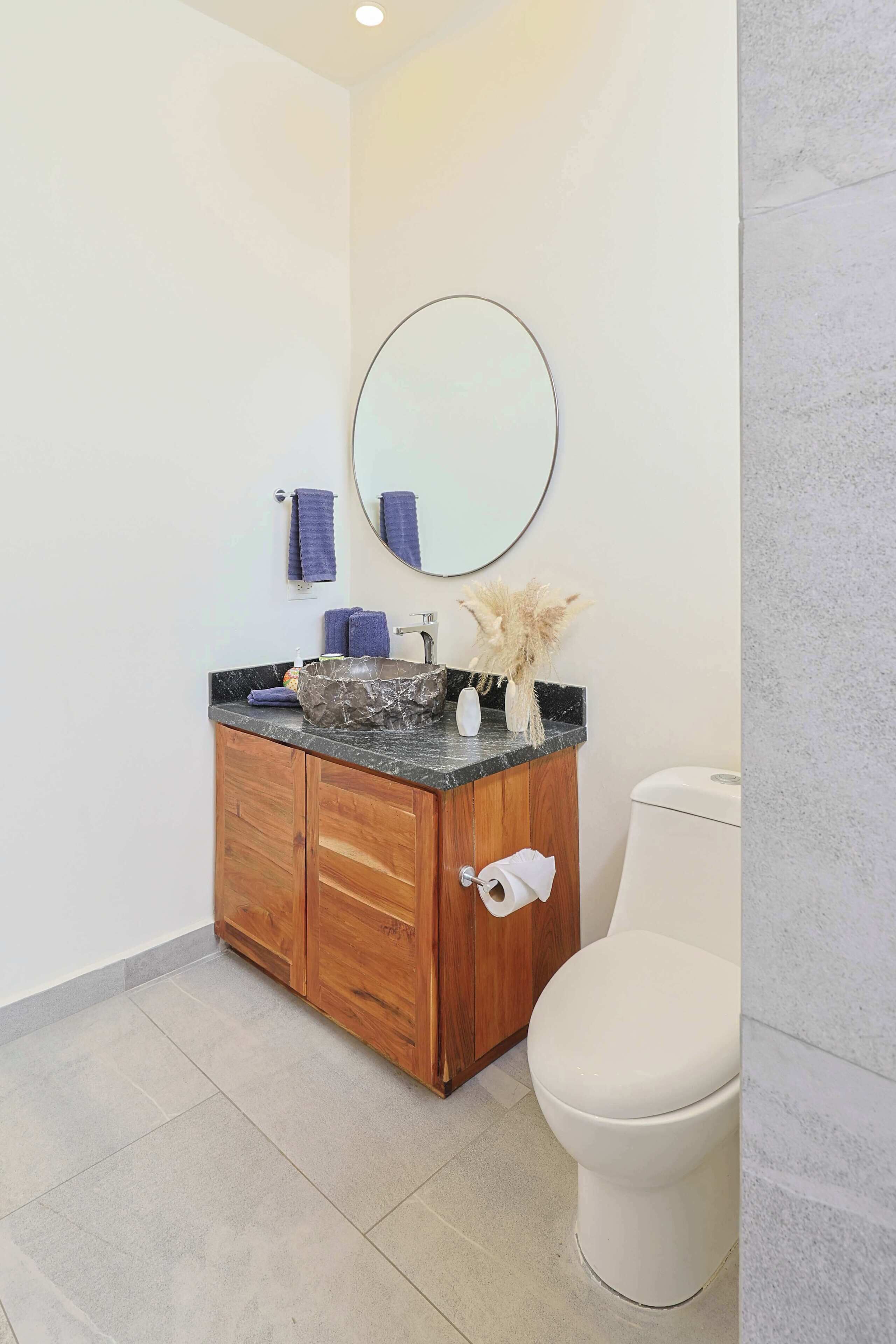 A bathroom with a wooden vanity, a round mirror, a white toilet, and a tiled floor.