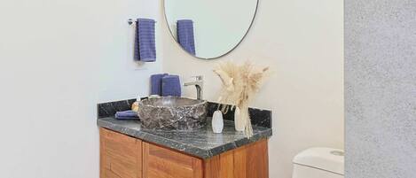 A bathroom with a wooden vanity, a round mirror, a white toilet, and a tiled floor.