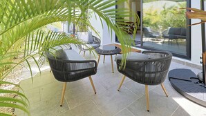 Un área de estar moderna al aire libre con sillas de mimbre y una mesa redonda, rodeada de grandes ventanales y plantas en macetas.