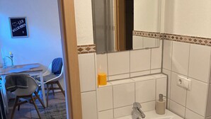 Un baño con lavamanos, espejo e inodoro. En la habitación contigua, hay una silla y una mesa con un florero.