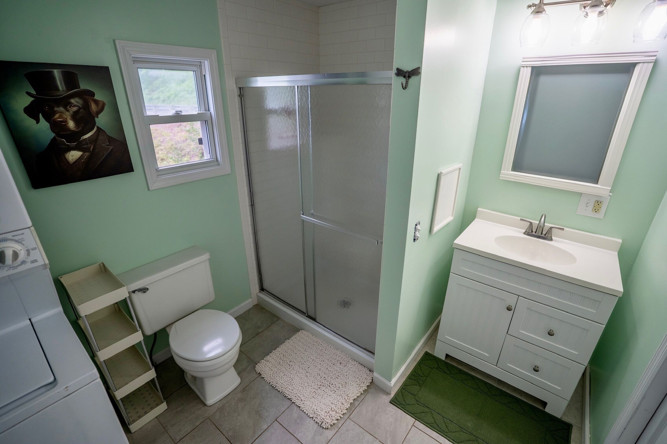 A bathroom with a toilet, shower, sink, and a painting of a dog.