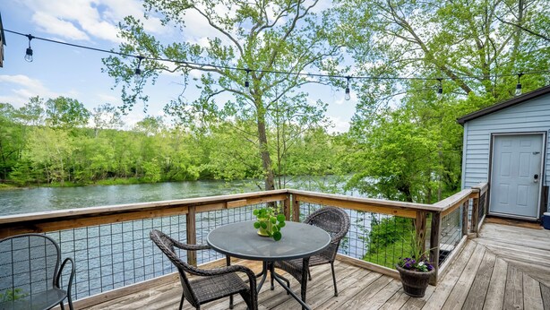 Een houten terras met een ronde tafel en stoelen, met uitzicht op een rivier en omringd door groen.