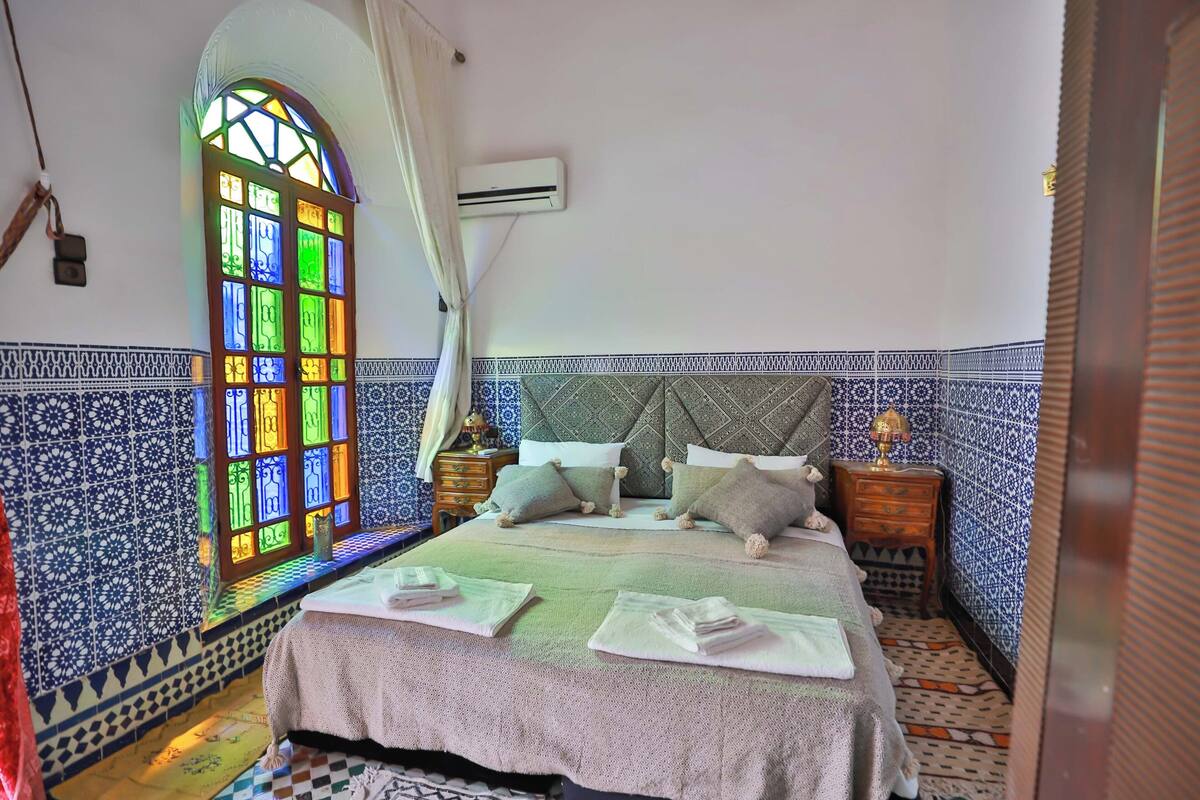 A neatly made bed with pillows and a blanket, a bedside table with a lamp, and a colorful stained glass window.