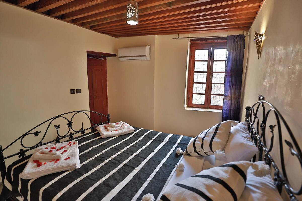 A bedroom with a bed, a window with curtains, an air conditioning unit, and a wooden ceiling.