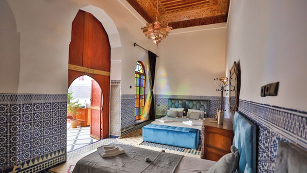 Ein Zimmer mit einem Bett, einem blauen Ottomanen, einer Holztür und einem Buntglasfenster.