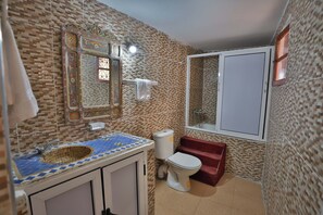 Et badeværelse med væg af mønstret fliseværk, en vask med bordplade i blåt og gult mosaik, et hvidt toilet og et spejl med et detaljeret rammeværk.