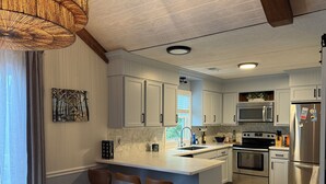 A modern kitchen with white cabinets, a central island with stools, stainless steel appliances, and a hanging light fixture. - Gorgeous Home on Sunset Beach, Just Steps from the Shore! (Sunset Beach)