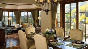 A dining room with a large table set for six, surrounded by six chairs. The room has large windows with a view of a forested area.