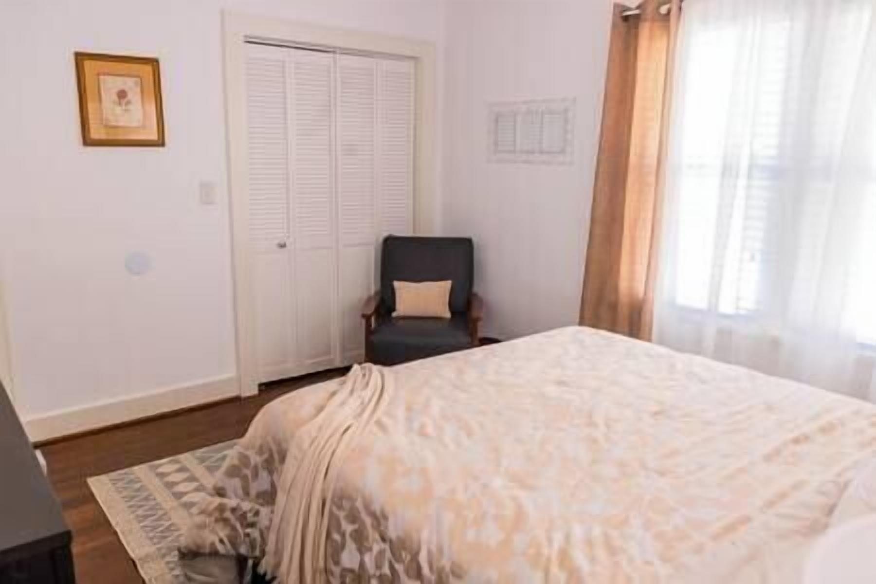 Un dormitorio con cama, una silla, armario y ventana con cortinas.