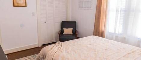Un dormitorio con cama, una silla, armario y ventana con cortinas.