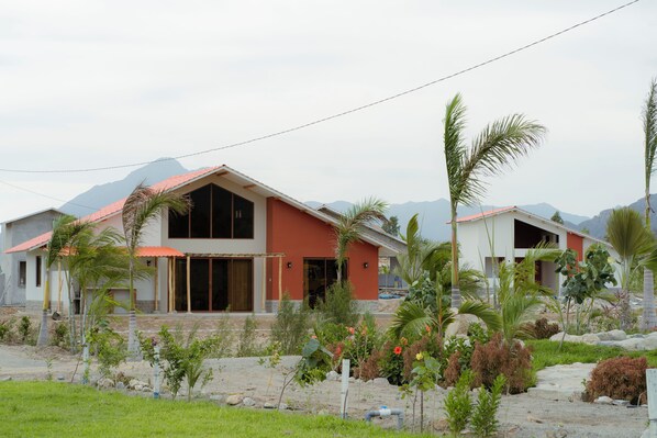 Una casa moderna de dos pisos con techo rojo, rodeada de plantas tropicales y un paisaje montañoso.