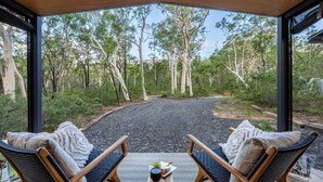 A covered outdoor seating area with two wicker chairs, a table with a glass of red wine and a plate of food, and a view of a forested area with a dirt path and a bench in the distance. - The Hollows - Kangaroo Valley (Kangaroo Valley)