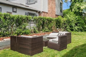 An outdoor patio with wicker furniture and a glass table, surrounded by well-maintained hedges and a brick wall.