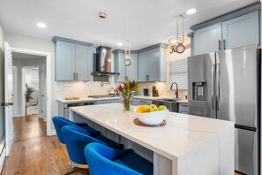A modern kitchen with grey cabinets, a stainless steel refrigerator, and a central island with seating. - Stylish 3BR Near NYC Train Parking + EV Charger (Maplewood)
