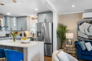 A modern kitchen with a central island, stainless steel appliances, and a dining area with blue chairs. - Stylish 3BR Near NYC Train Parking + EV Charger (Maplewood)