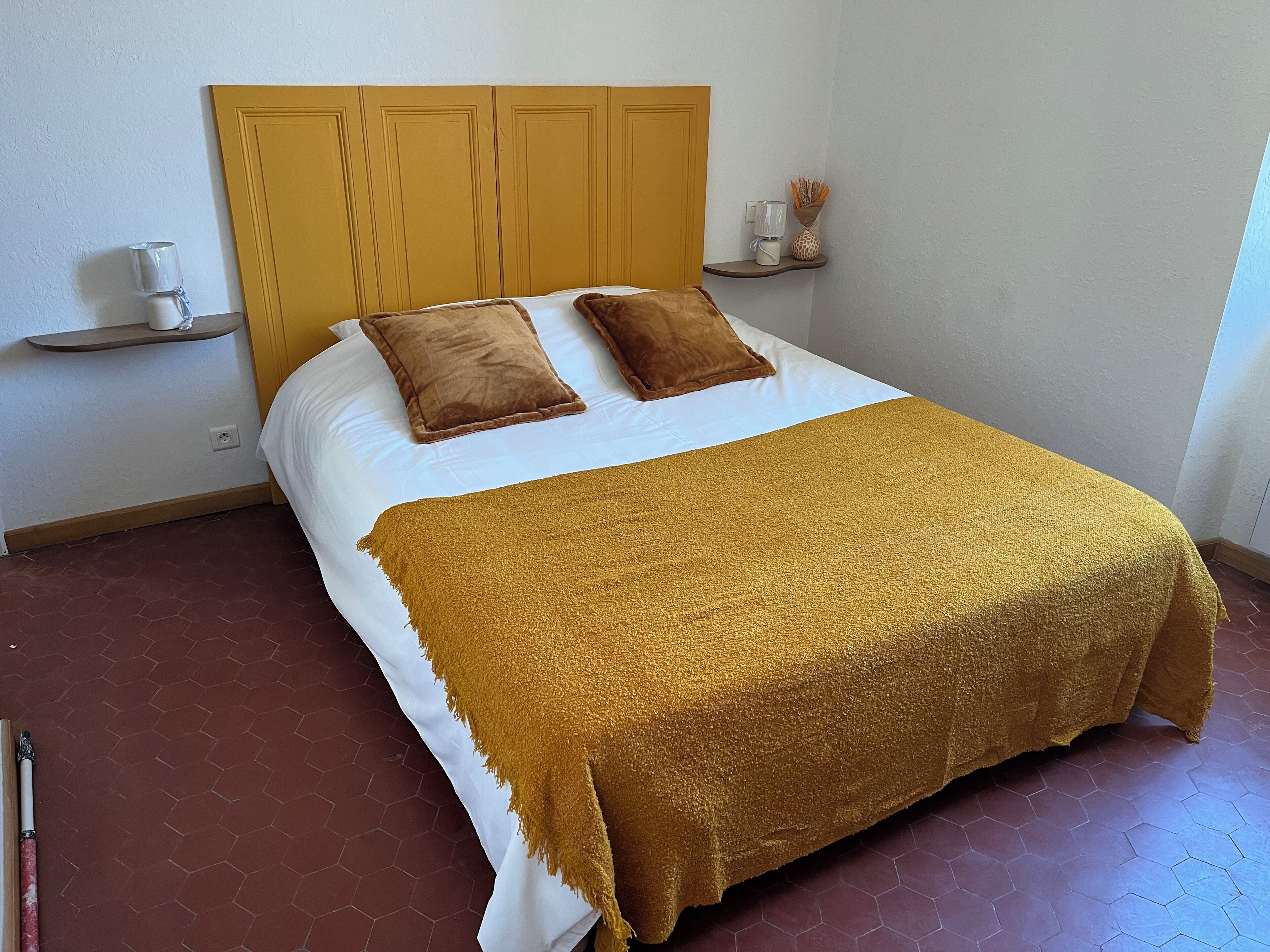 Una cama bien hecha con un edredón marrón y dos almohadas, cabecera de madera, un estante con una taza y un adorno, y piso de baldosas.