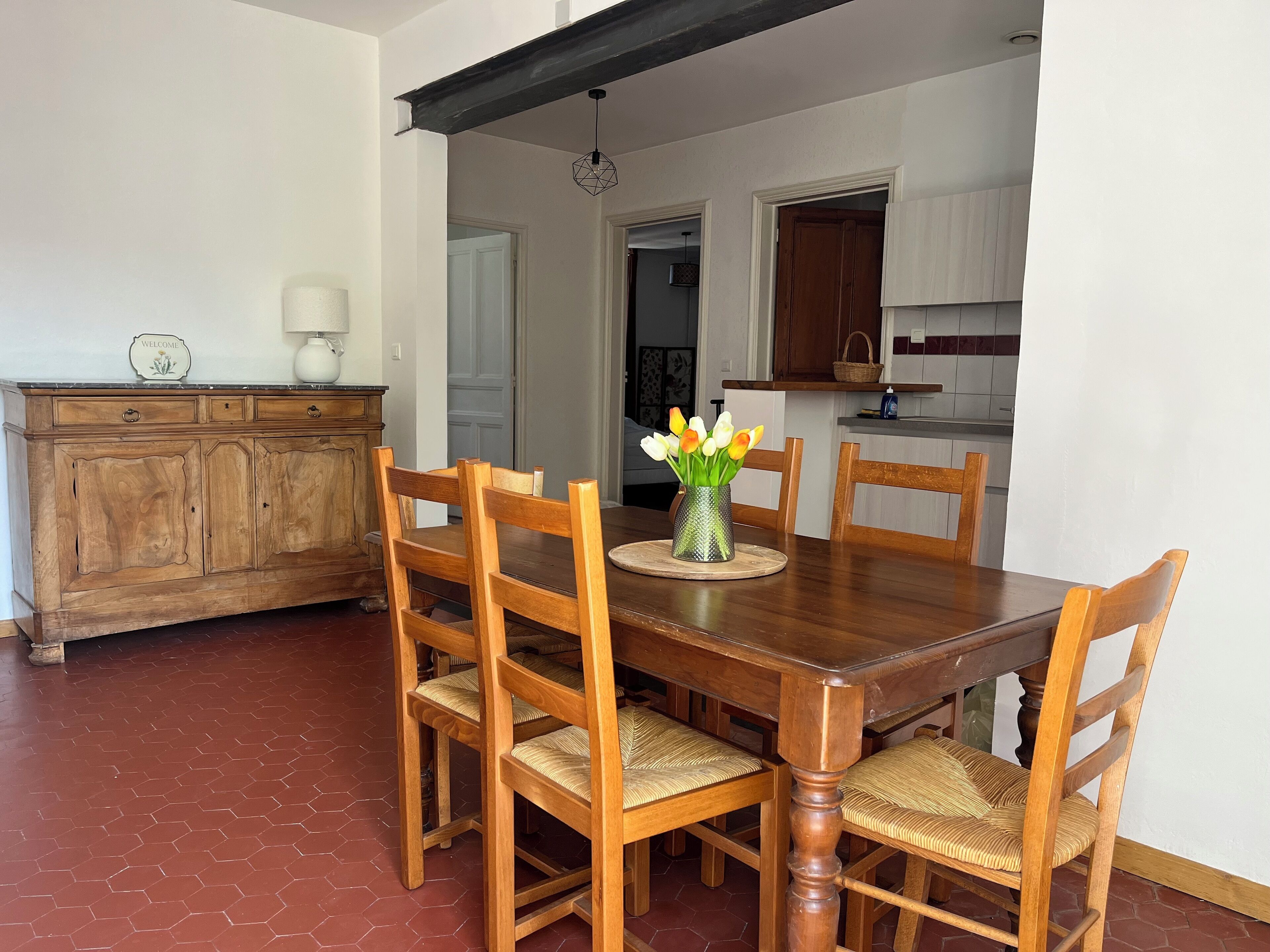 Un'area pranzo con tavolo e sedie in legno, un vaso con fiori e una cucina sullo sfondo.
