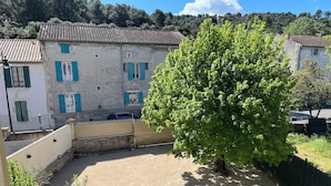 A courtyard with a large tree, a building with blue shutters, and a stone wall. - House in the village, near Avignon (Barbentane)