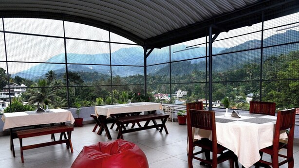 Un espace extérieur abrité, aménagé pour les repas, avec des tables et des chaises, un pouf rouge et une vue sur les montagnes et la verdure.