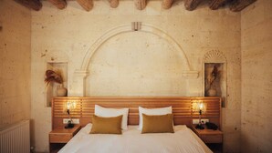 A bedroom with a wooden headboard, two bedside tables with lamps, a large bed with white bedding, and a stone wall with a decorative arch. - Juno Cappadocia (Nevsehir)