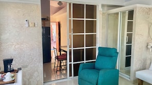 A room with a teal chair, a sliding glass door, a coffee maker, and a dining area visible through the door.
