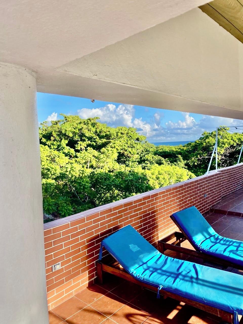Un balcón con dos sillones azules, un muro de ladrillo y vistas a árboles verdes y un cielo despejado.