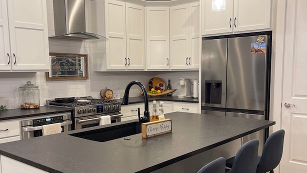Una cocina moderna con gabinetes blancos, electrodomésticos de acero inoxidable, una isla central con taburetes y luminarias colgantes.