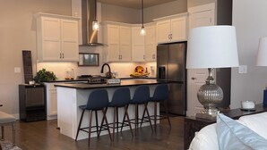 Cocina moderna con armarios blancos, una isla central con taburetes de bar, un refrigerador de acero inoxidable e iluminación colgante.