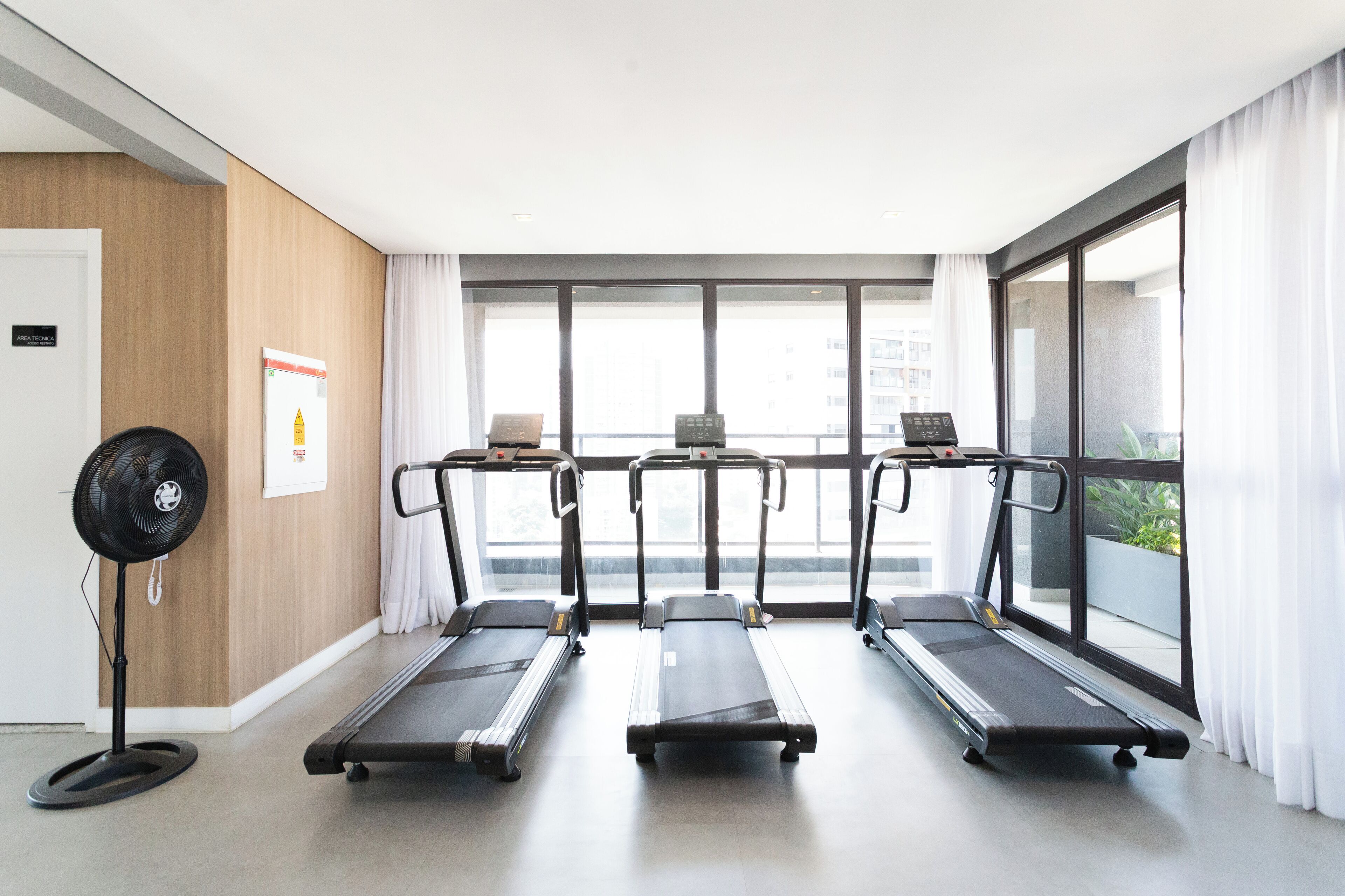 A gym with three treadmills, a standing fan, and large windows with curtains.