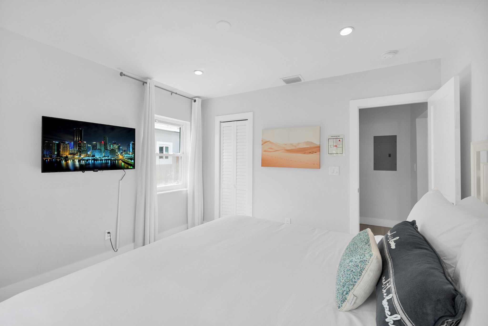 A modern hotel room with a large bed, a cityscape painting, a desert landscape painting, and a door leading to another room.