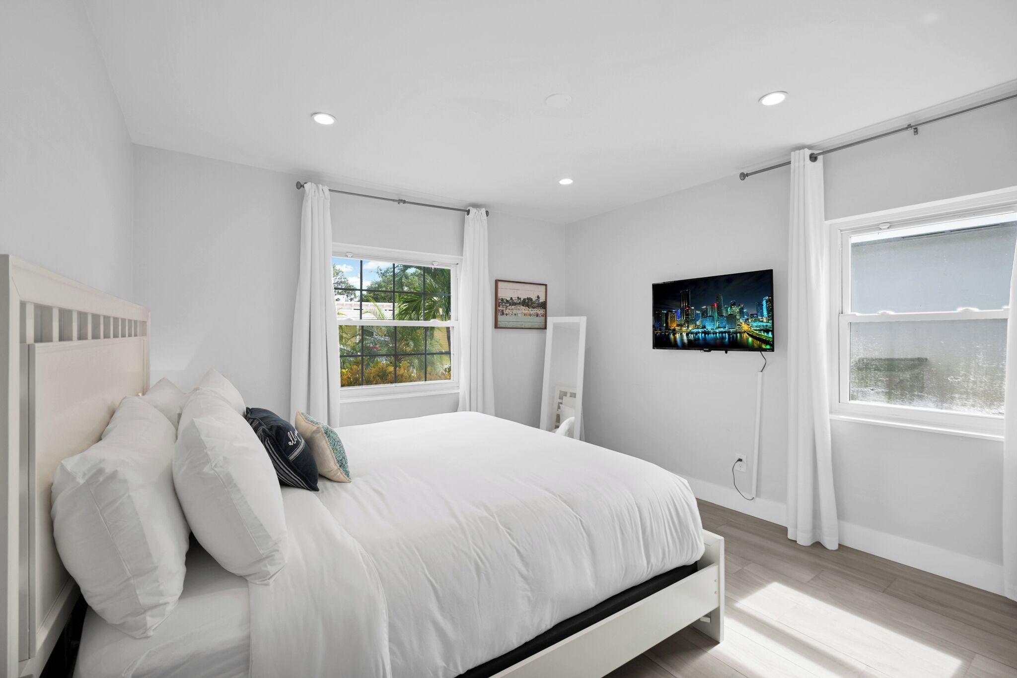 Une chambre moderne avec un grand lit, des rideaux blancs, un téléviseur fixé au mur et une fenêtre donnant sur un panorama urbain.