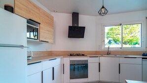 A modern kitchen with white cabinets, a built-in oven, a gas stove, and a window with a view of trees. - Le Narbon - maison proche de la plage (Erdeven)