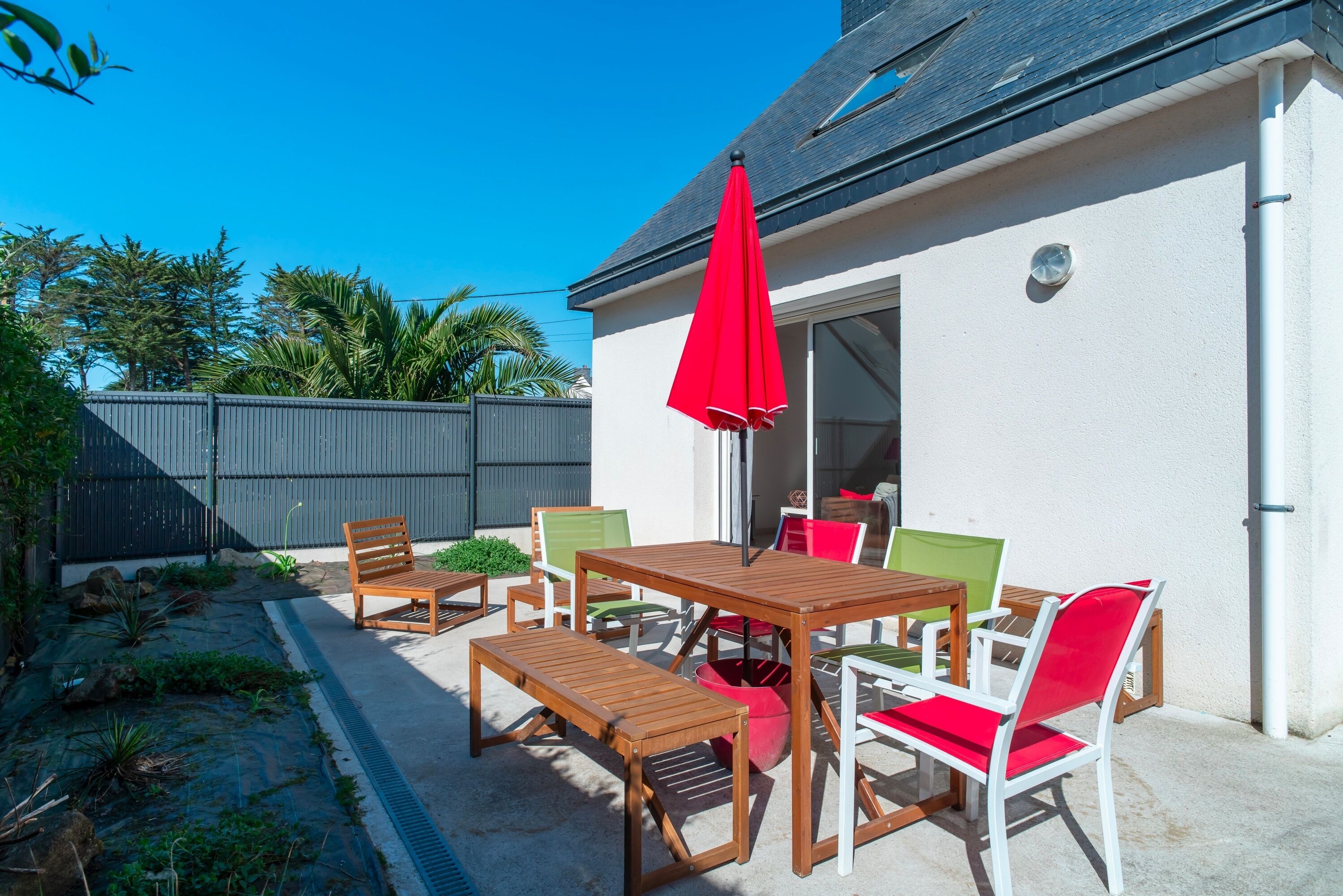 A patio with wooden furniture and a red umbrella, a white wall, and a grey fence.