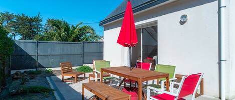 A patio with wooden furniture and a red umbrella, a white wall, and a grey fence.