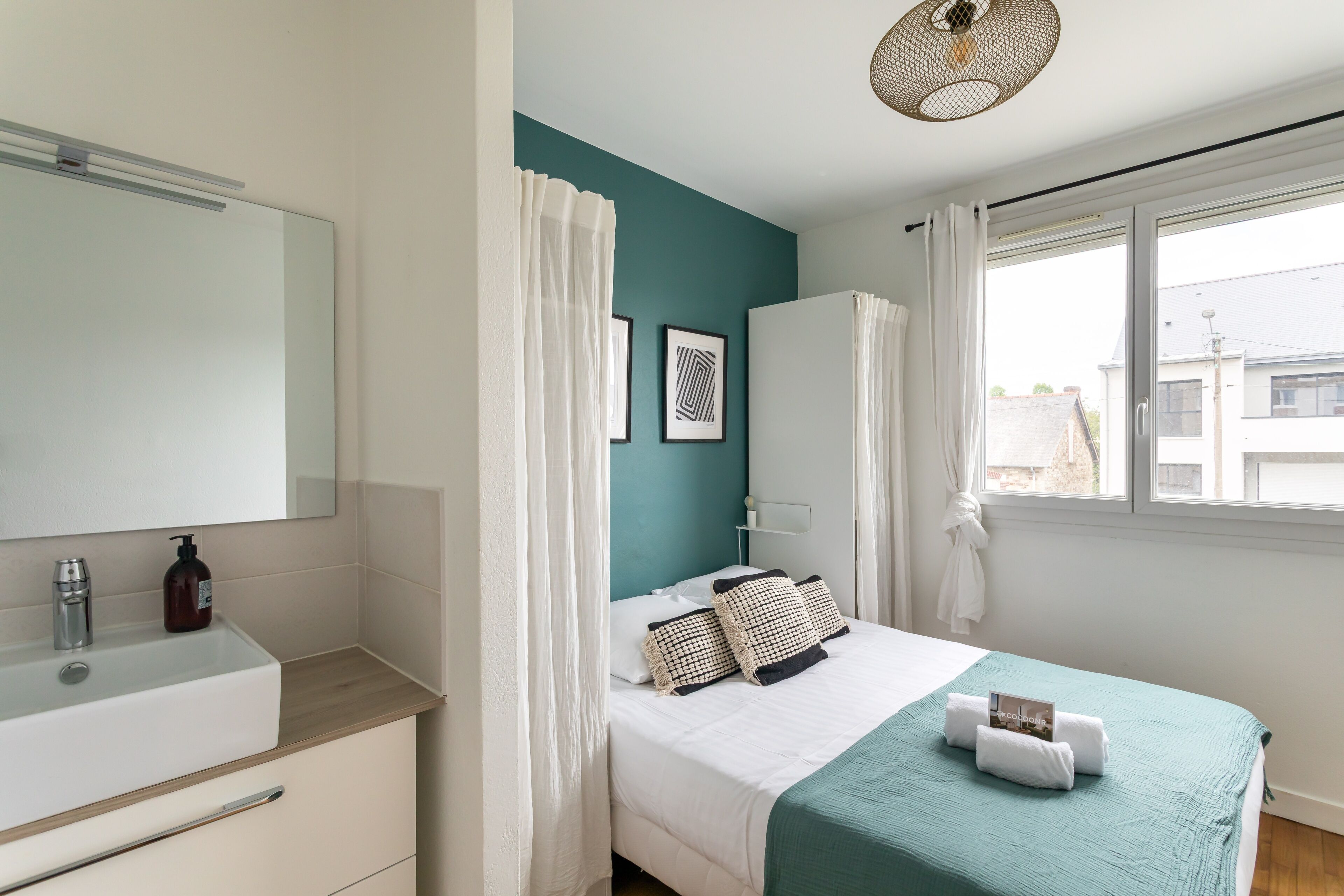Une chambre moderne avec un mur d’accent de couleur turquoise, un grand lit avec une literie blanche, une armoire blanche et une fenêtre avec des rideaux.