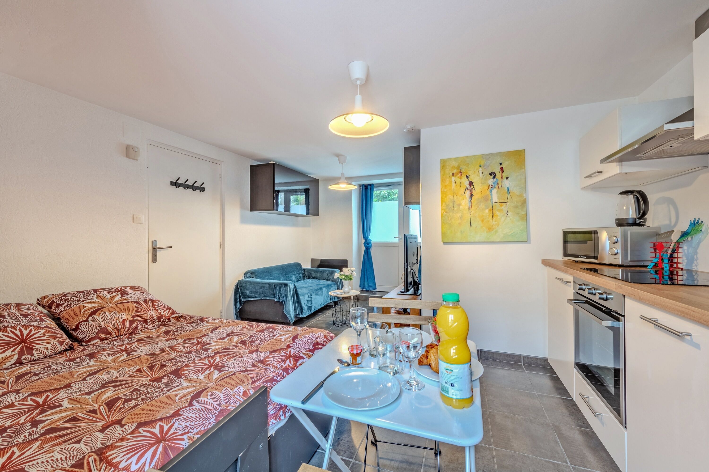 A compact living space with a bed, a dining table set with plates and glasses, a kitchen area with a microwave and kettle, and a blue chair.