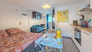 A compact living space with a bed, a dining table set with plates and glasses, a kitchen area with a microwave and kettle, and a blue chair.