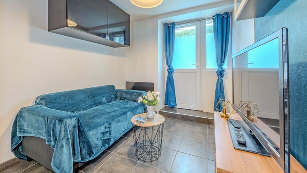 A modern room with a blue sofa, a small round table with a vase of flowers, a flat-screen TV, and a window with blue curtains.