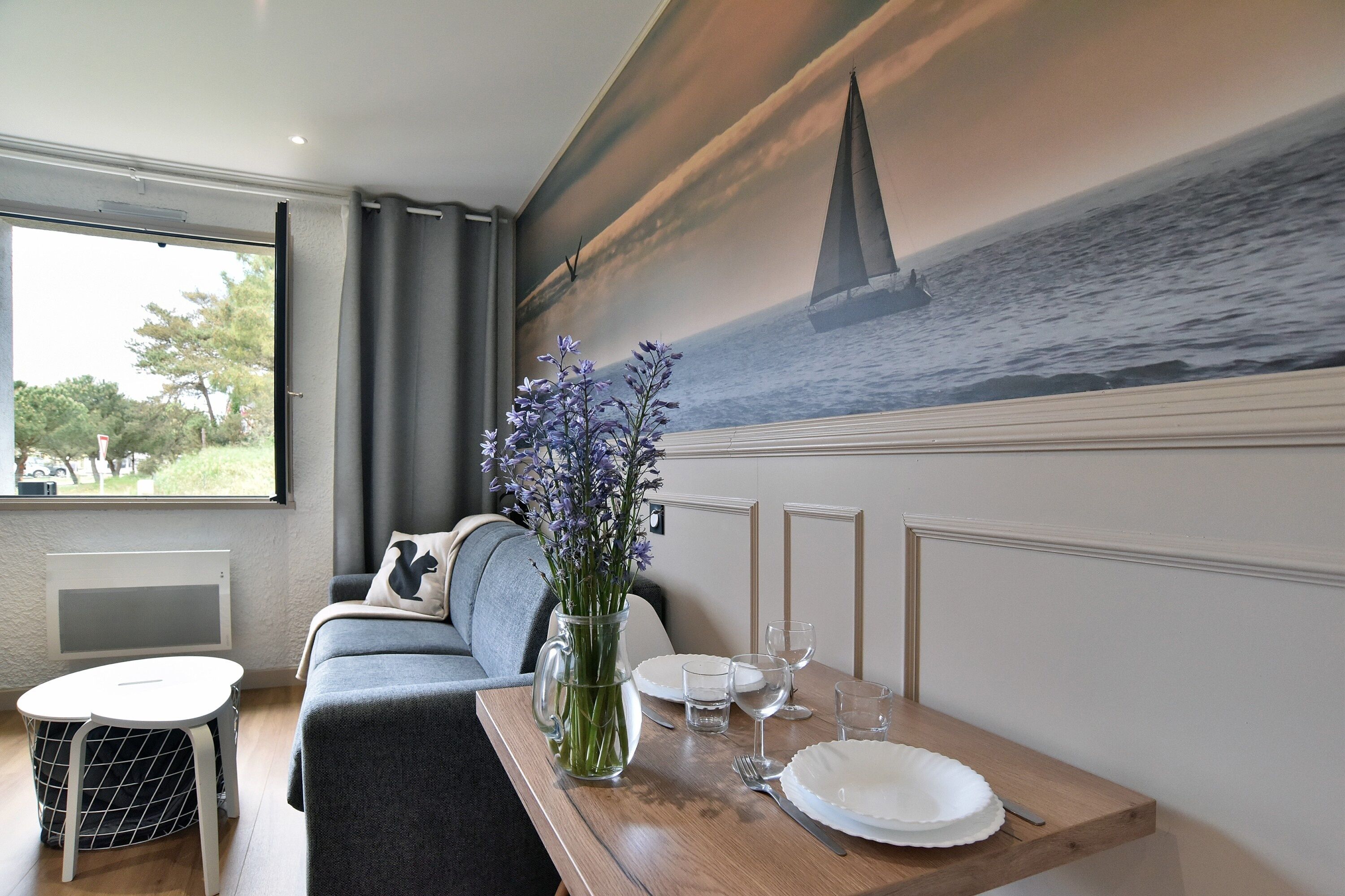 Een eetruimte met een houten tafel voor twee personen, een bloemenvaas en een muurschildering van een zeilboot.