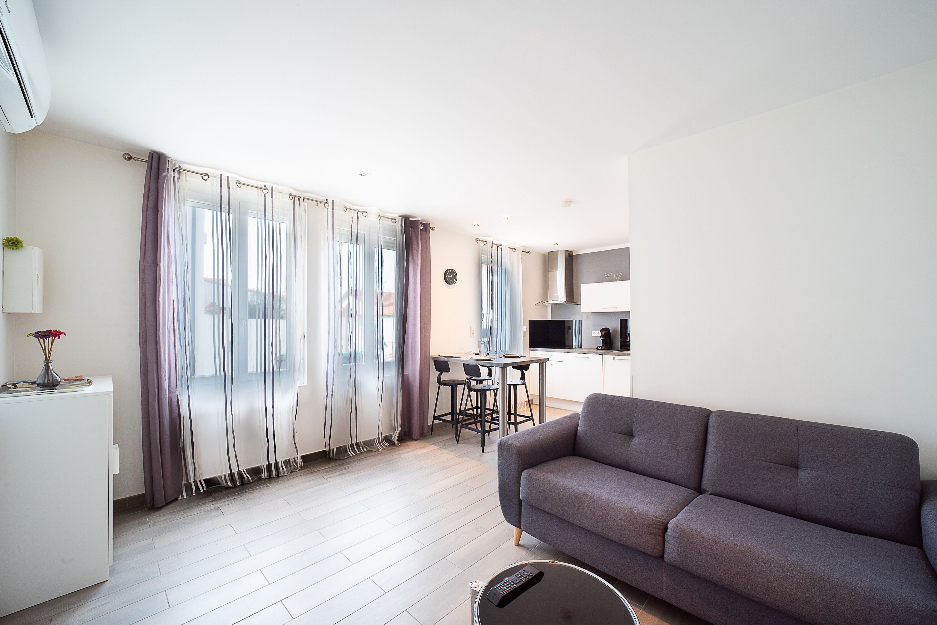 Ein modernes Wohnzimmer mit grauem Sofa, einem kleinen weißen Tisch und einer Küchenzeile mit Fernseher und Mikrowelle.