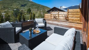 Una terraza de madera con mobiliario exterior, una mesa de vidrio y vista a la montaña.