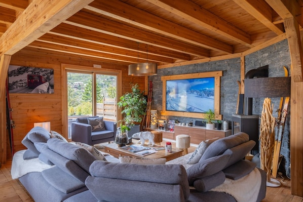 A cozy cabin interior with a fireplace, wooden beams, a large window, and a sofa.