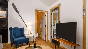A modern living room with a blue armchair, a round wooden coffee table, a TV on a wooden stand, and a lamp on a small table. - Lakeview Deck| 10 mi from TC | Sauna | Hot Tub (Traverse City)