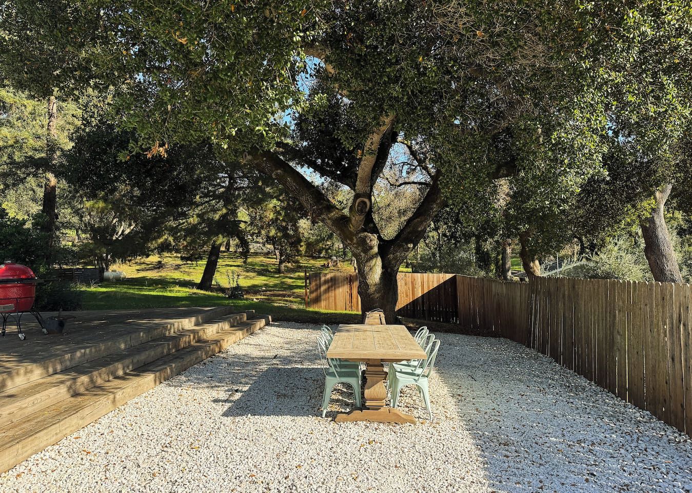 Un patio exterior con una mesa y sillas de madera, rodeado de grava y árboles.