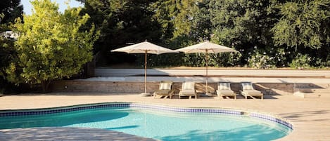 Piscina con cubierta de madera, rodeada de árboles y equipada con sillones de descanso y sombrillas.