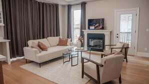 Een woonkamer met een bank, twee fauteuils, een glazen salontafel, een open haard en een televisie.