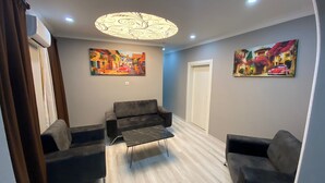 Ein modernes Wohnzimmer mit grauem Sofa, einem schwarzen Couchtisch und zwei Sesseln. An den Wänden hängen zwei Bilder, eine Deckenleuchte und eine Klimaanlage.
