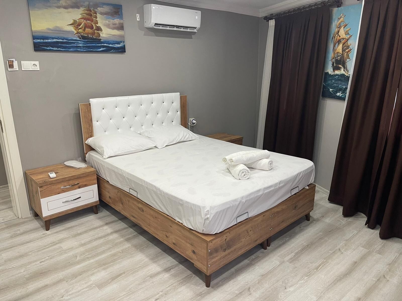 Una cama bien hecha con sábanas blancas y toallas enrolladas, una mesita de noche de madera con cajón, un aire acondicionado instalado en la pared y dos cuadros que representan barcos en el mar.